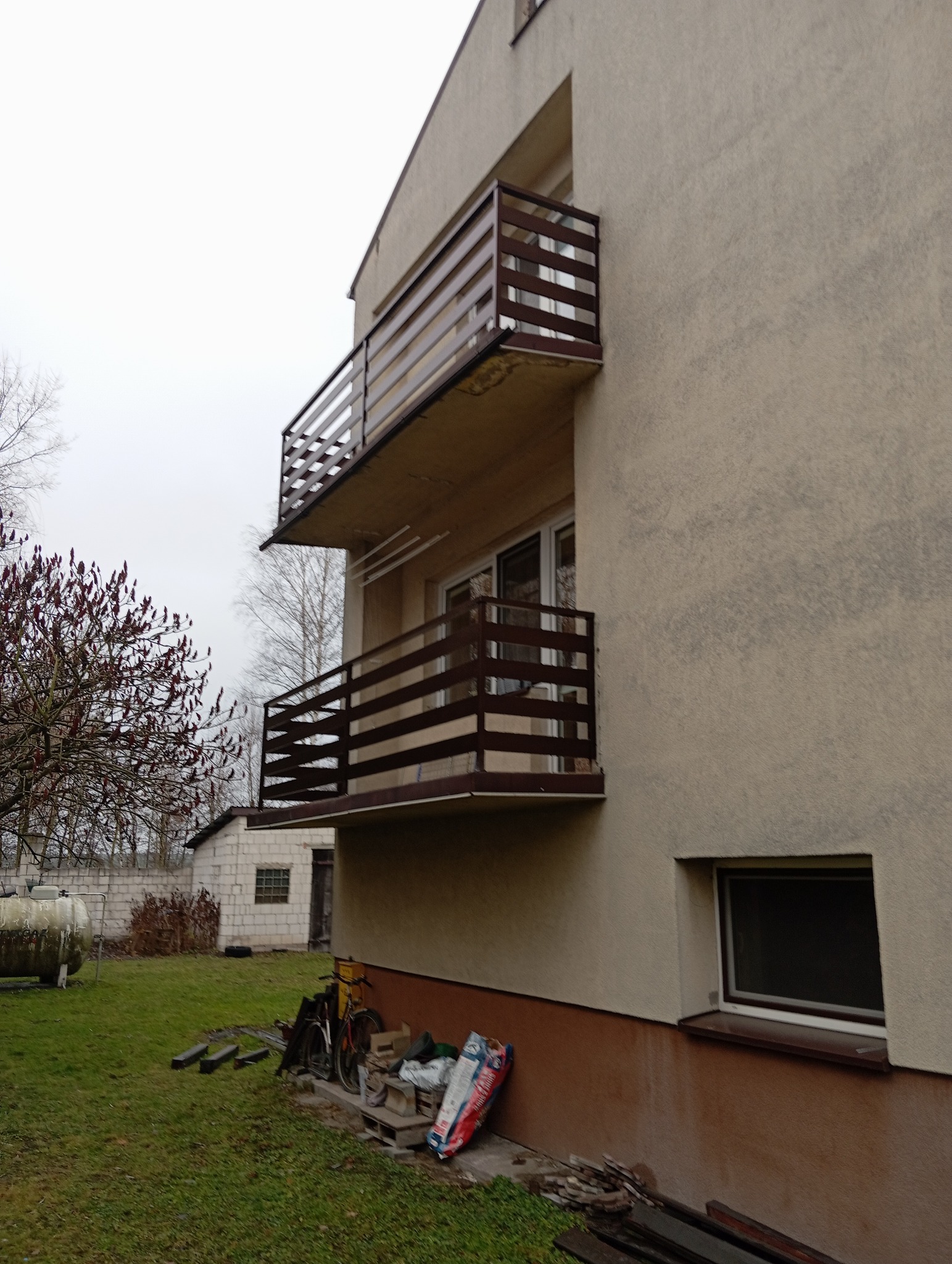 Dwukondygnacyjny budynek z drewnianymi balustradami balkonowymi o poziomej konstrukcji, widok z boku, na trawniku rowery i materiały budowlane.