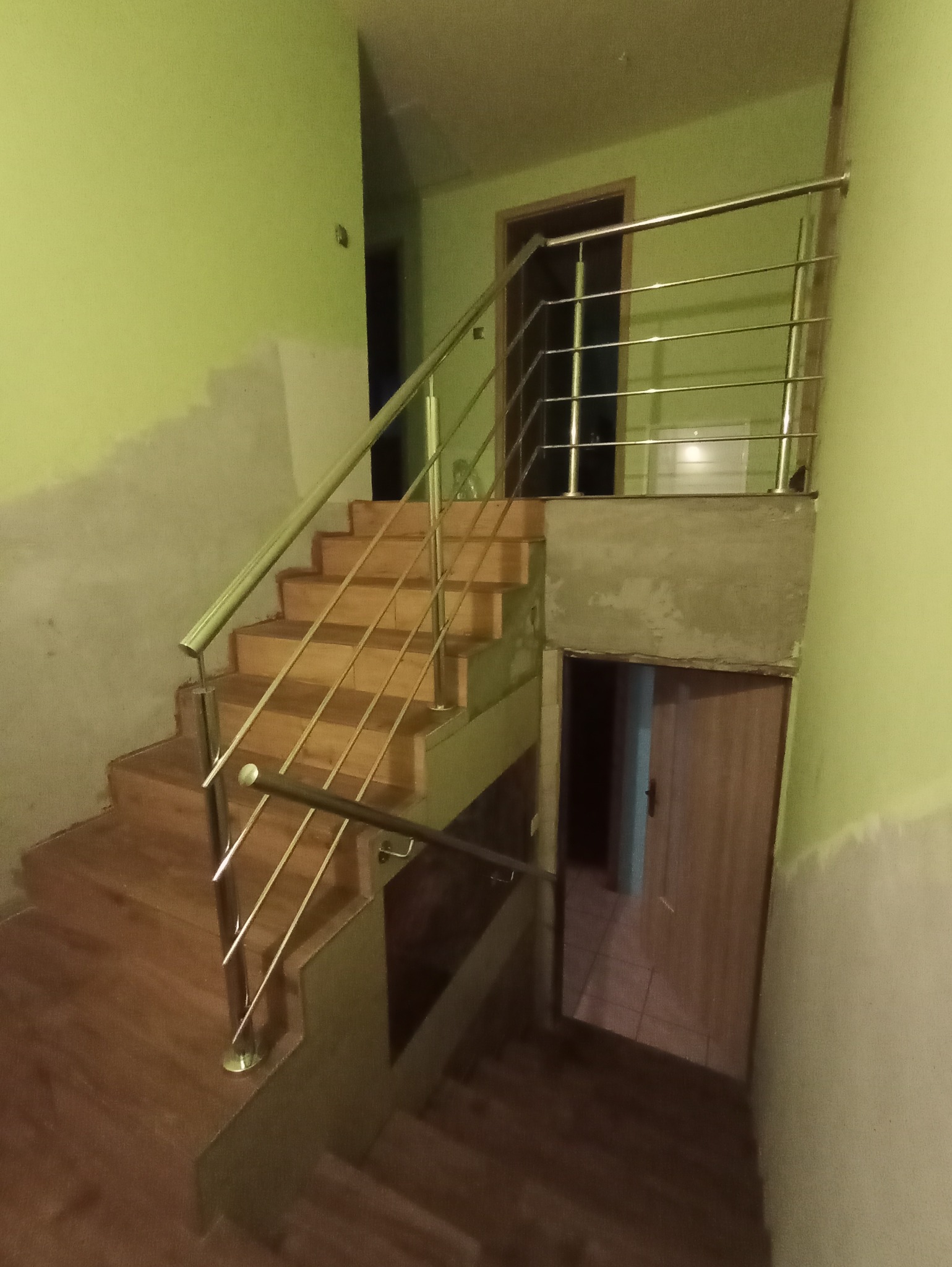 Drewniane schody wewnętrzne z balustradą ze stali nierdzewnej, widok z dołu, ściany w kolorze pistacjowym, widoczne niedokończone wykończenia ścian.