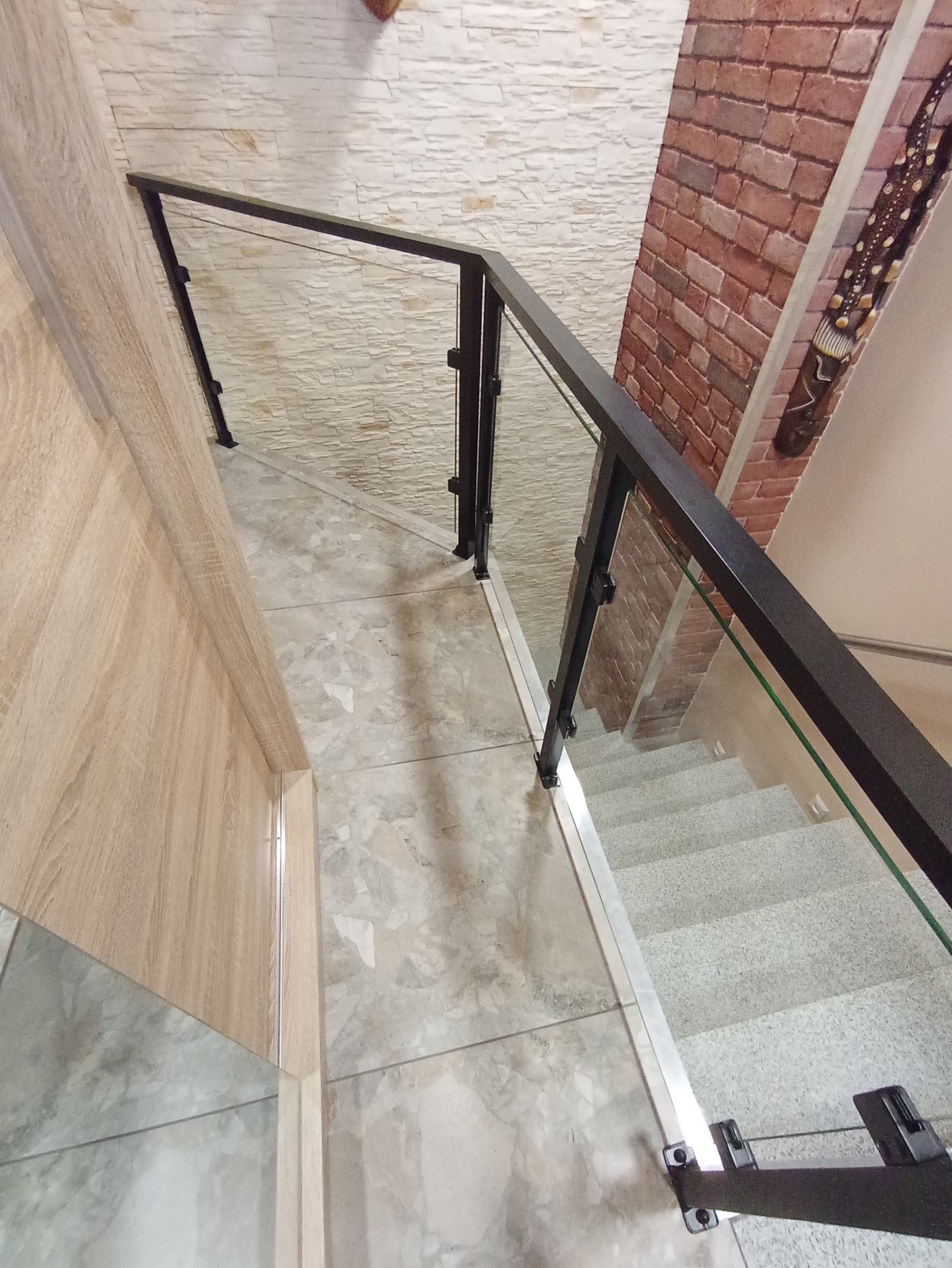 Widok z góry na schody z szarymi stopniami i szklaną balustradą z czarnymi elementami mocującymi, ściany wykończone w różnych stylach: cegła, kamień i drewno.