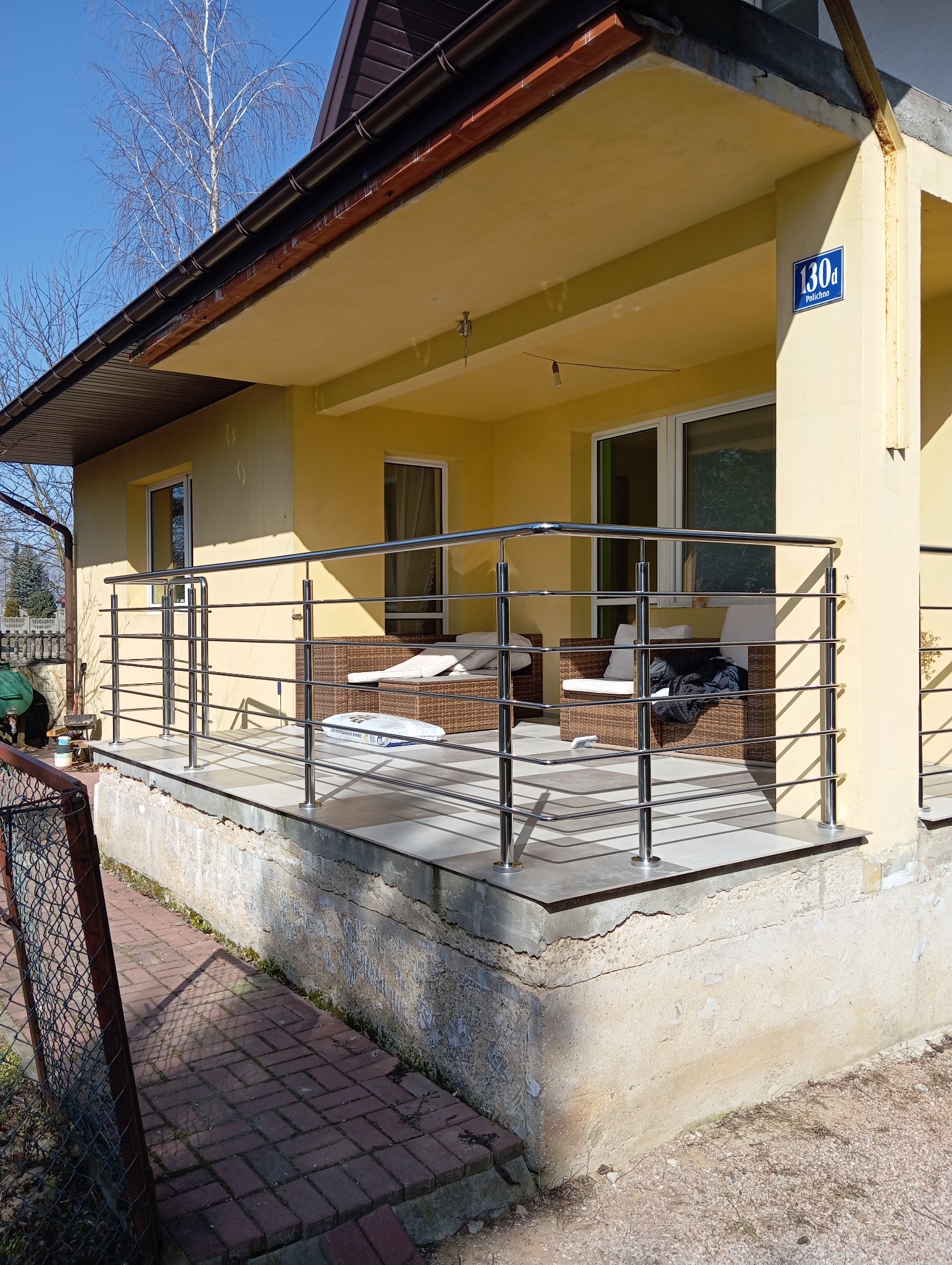 Balkon z balustradą ze stali nierdzewnej przy żółtym domu z numerem 130d, widok z zewnątrz, na balkonie rattanowe meble.