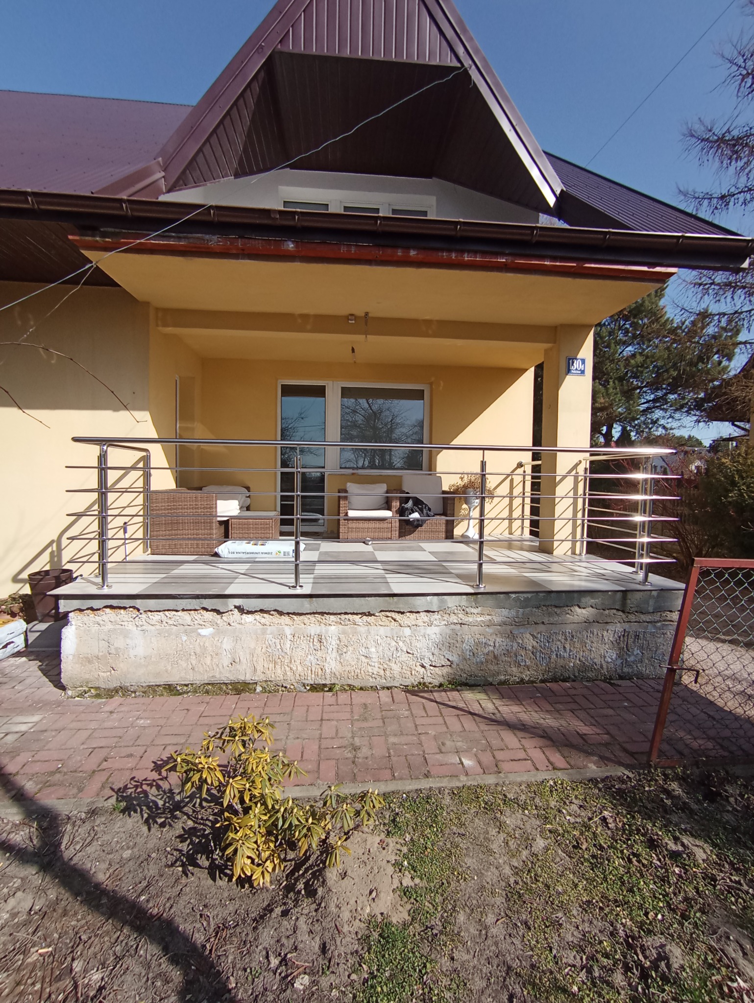 Zewnętrzny taras domu z chromowanymi balustradami, rattanowymi meblami wypoczynkowymi i płytkami w szachownicę.