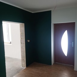 Usługi remontowo- wykończeniowe - Pomieszczenie po remoncie: ściany pomalowane w kolorze butelkowej zieleni, podłoga wyłożona panelami imitującymi drewno, widoczne drzwi z owalnym przeszkleniem i surowy otwór drzwiowy do kolejnego...