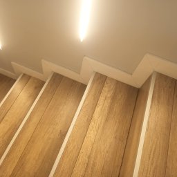 Usługi remontowo- wykończeniowe - Drewnopodobne panele na schodach z białymi listwami przypodłogowymi i oświetleniem LED w suficie.
