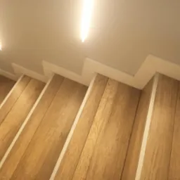 Drewnopodobne panele na schodach z białymi listwami przypodłogowymi i oświetleniem LED w suficie.