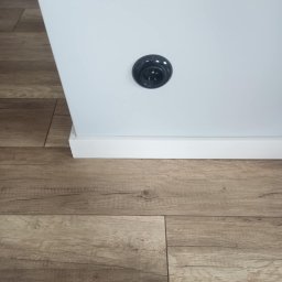 Usługi remontowo- wykończeniowe - Fragment ściany pomalowanej na biało z czarnym gniazdkiem elektrycznym oraz podłoga z paneli imitujących drewno.