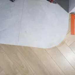 Połączenie szarej płytki ceramicznej i jasnych paneli ułożonych w jodełkę, widoczne narzędzia w rogu, wykończenie z białym cokolikiem.