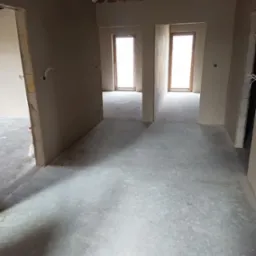 Wnętrze w stanie deweloperskim: surowe ściany z instalacją elektryczną, betonowa posadzka, widoczne drewniane elementy konstrukcji dachu, okna w drewnianych ramach w głębi pomieszczenia.