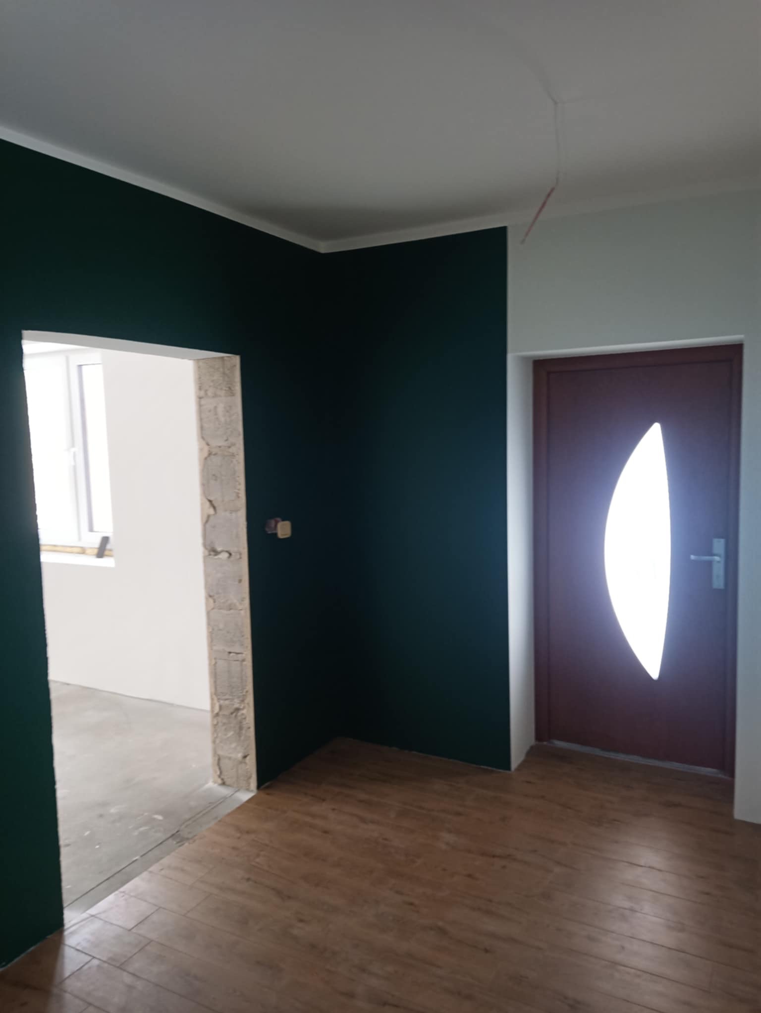 Pomieszczenie po remoncie: ściany pomalowane w kolorze butelkowej zieleni, podłoga wyłożona panelami imitującymi drewno, widoczne drzwi z owalnym przeszkleniem i surowy otwór drzwiowy do kolejnego...