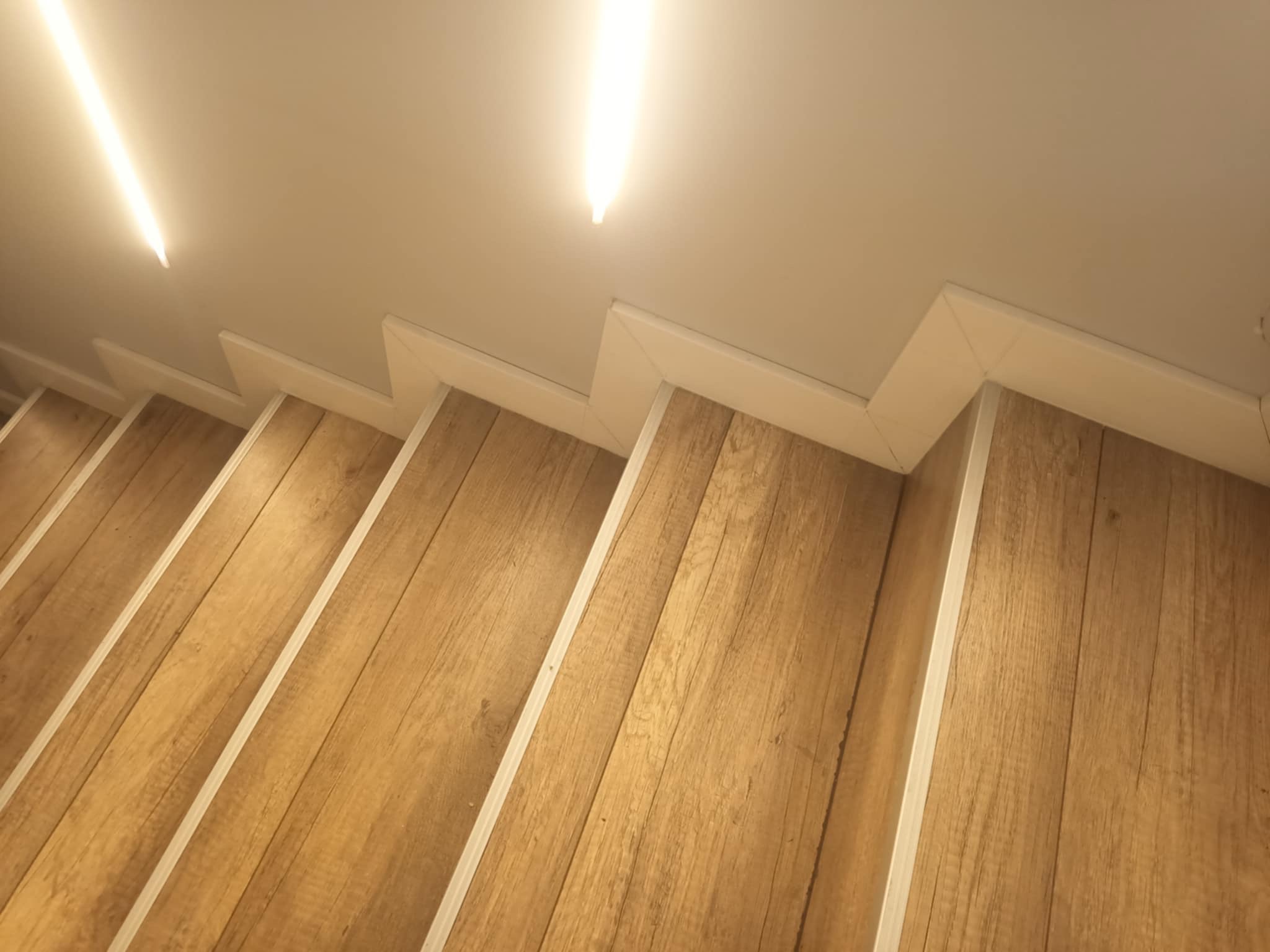 Drewnopodobne panele na schodach z białymi listwami przypodłogowymi i oświetleniem LED w suficie.