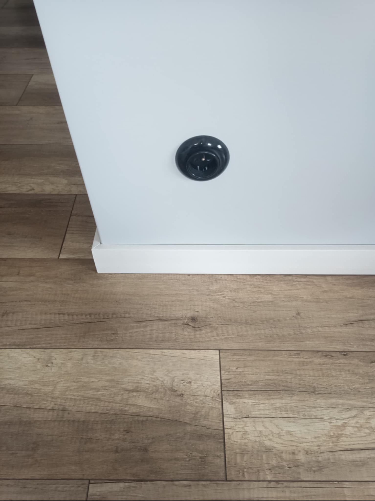 Fragment ściany pomalowanej na biało z czarnym gniazdkiem elektrycznym oraz podłoga z paneli imitujących drewno.