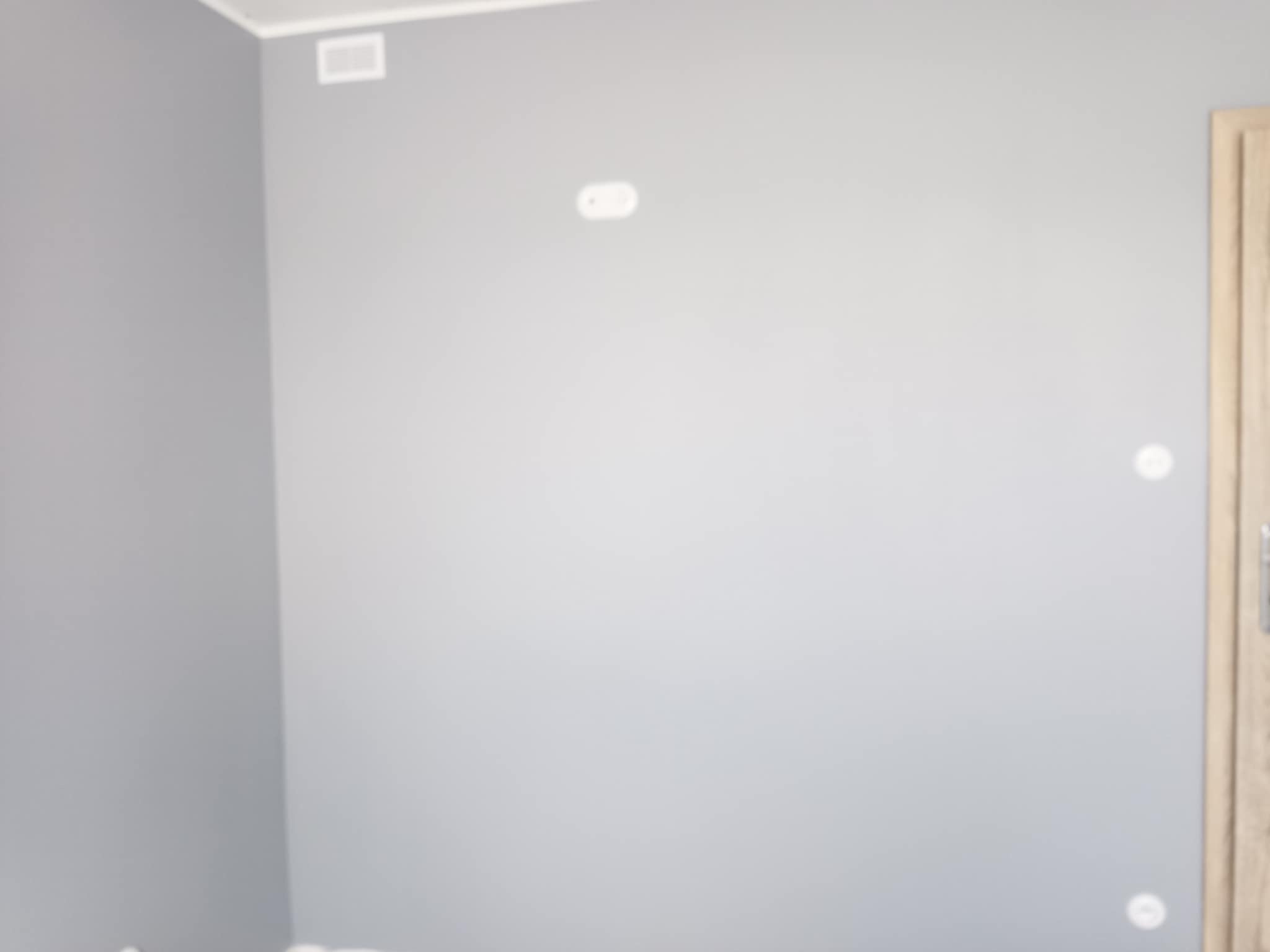 Świeżo pomalowane na szaro ściany w pokoju, widoczne gniazdka elektryczne i fragment drewnianej ościeżnicy drzwiowej po prawej stronie.