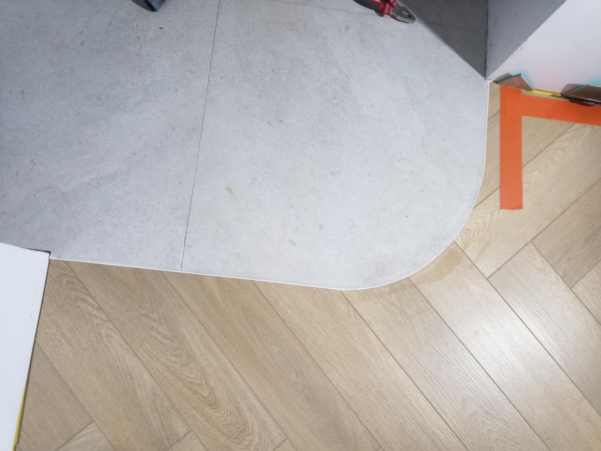 Połączenie szarej płytki ceramicznej i jasnych paneli ułożonych w jodełkę, widoczne narzędzia w rogu, wykończenie z białym cokolikiem.