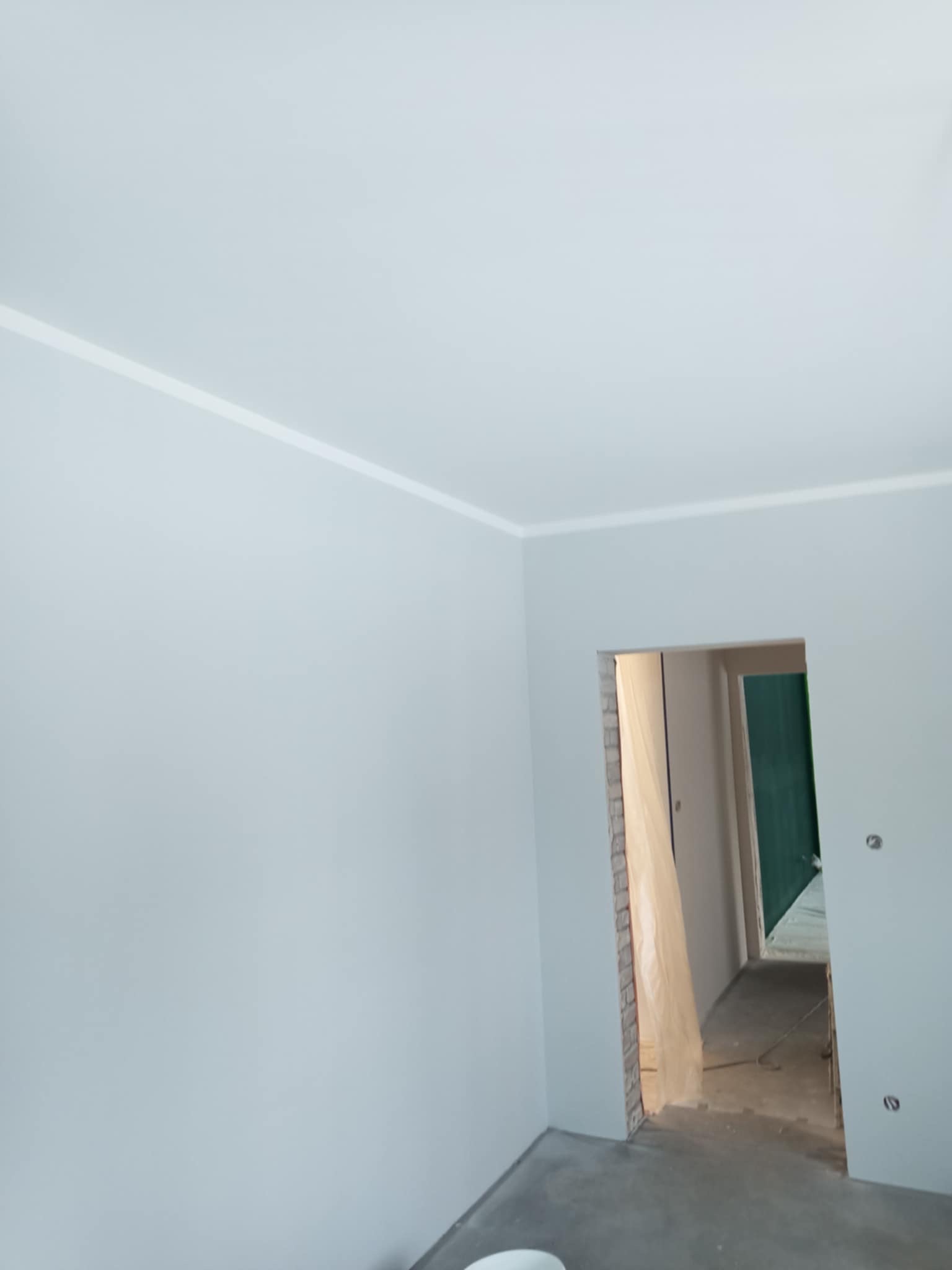 Świeżo pomalowane na szaro ściany i sufit z białym paskiem dekoracyjnym przy suficie w pokoju z otwartymi drzwiami do kolejnego pomieszczenia, w tle zabezpieczona folią ościeżnica.