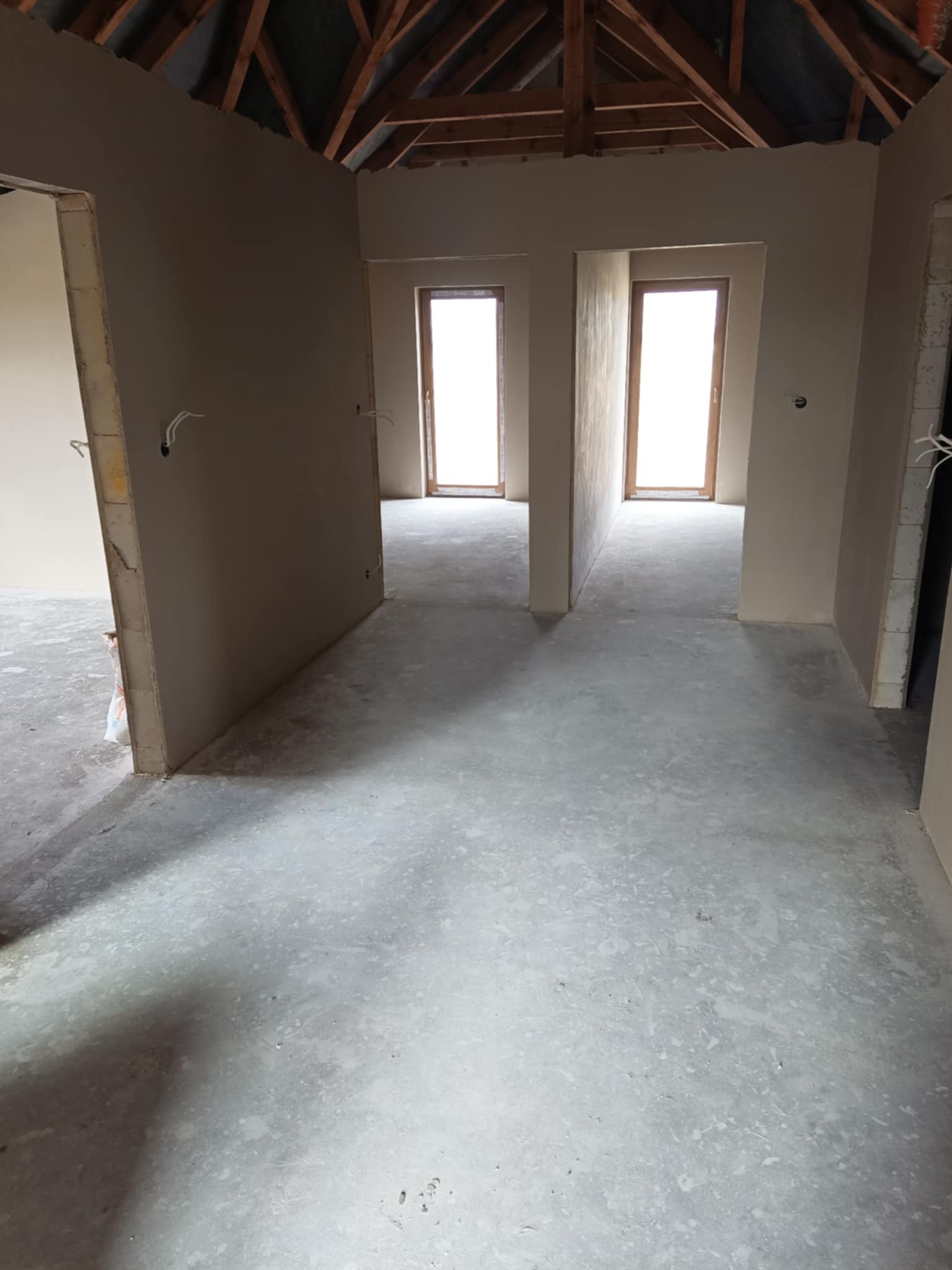 Wnętrze w stanie deweloperskim: surowe ściany z instalacją elektryczną, betonowa posadzka, widoczne drewniane elementy konstrukcji dachu, okna w drewnianych ramach w głębi pomieszczenia.