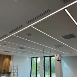 Nowoczesny sufit z oświetleniem liniowym LED i kratkami wentylacyjnymi w biurze. Widoczna drabina i fragment ściany z okładziną drewnopodobną.