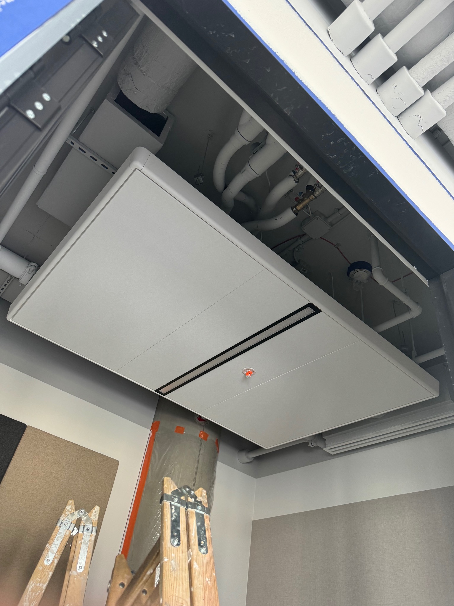 Montaż sufitu podwieszanego kasetonowego w pomieszczeniu biurowym. Widoczne rury i instalacje pod sufitem. Ustawiona drabina malarska.