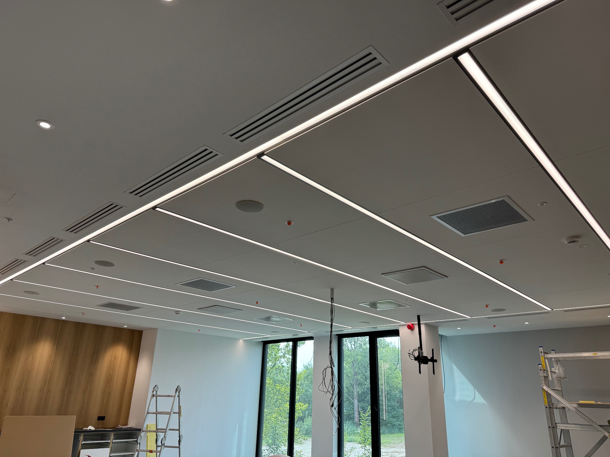 Nowoczesny sufit z oświetleniem liniowym LED i kratkami wentylacyjnymi w biurze. Widoczna drabina i fragment ściany z okładziną drewnopodobną.