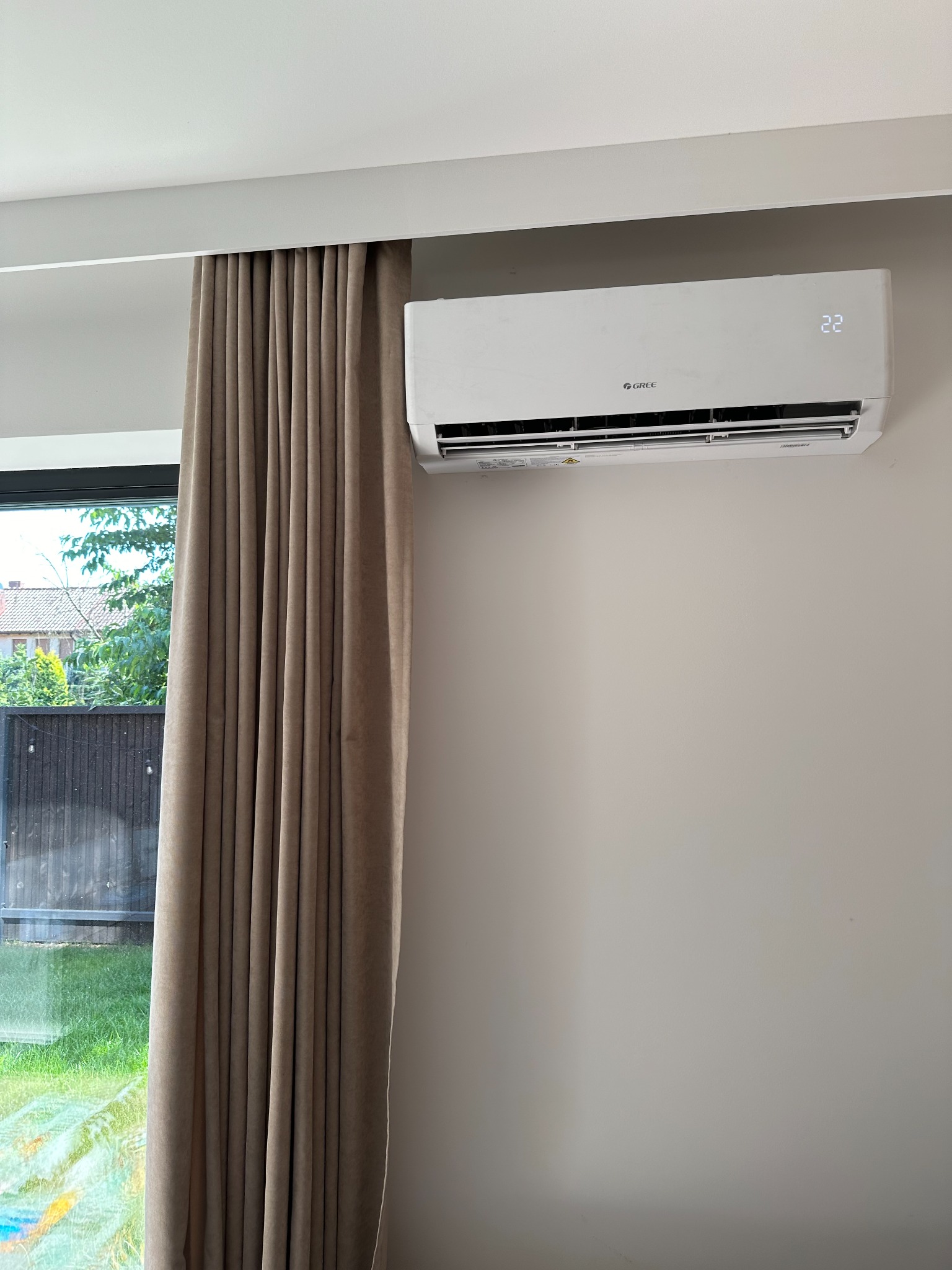 Biała jednostka klimatyzacyjna marki Gree zamontowana na ścianie w pokoju, obok okna z beżowymi zasłonami, wyświetlająca temperaturę 22 stopnie Celsjusza.