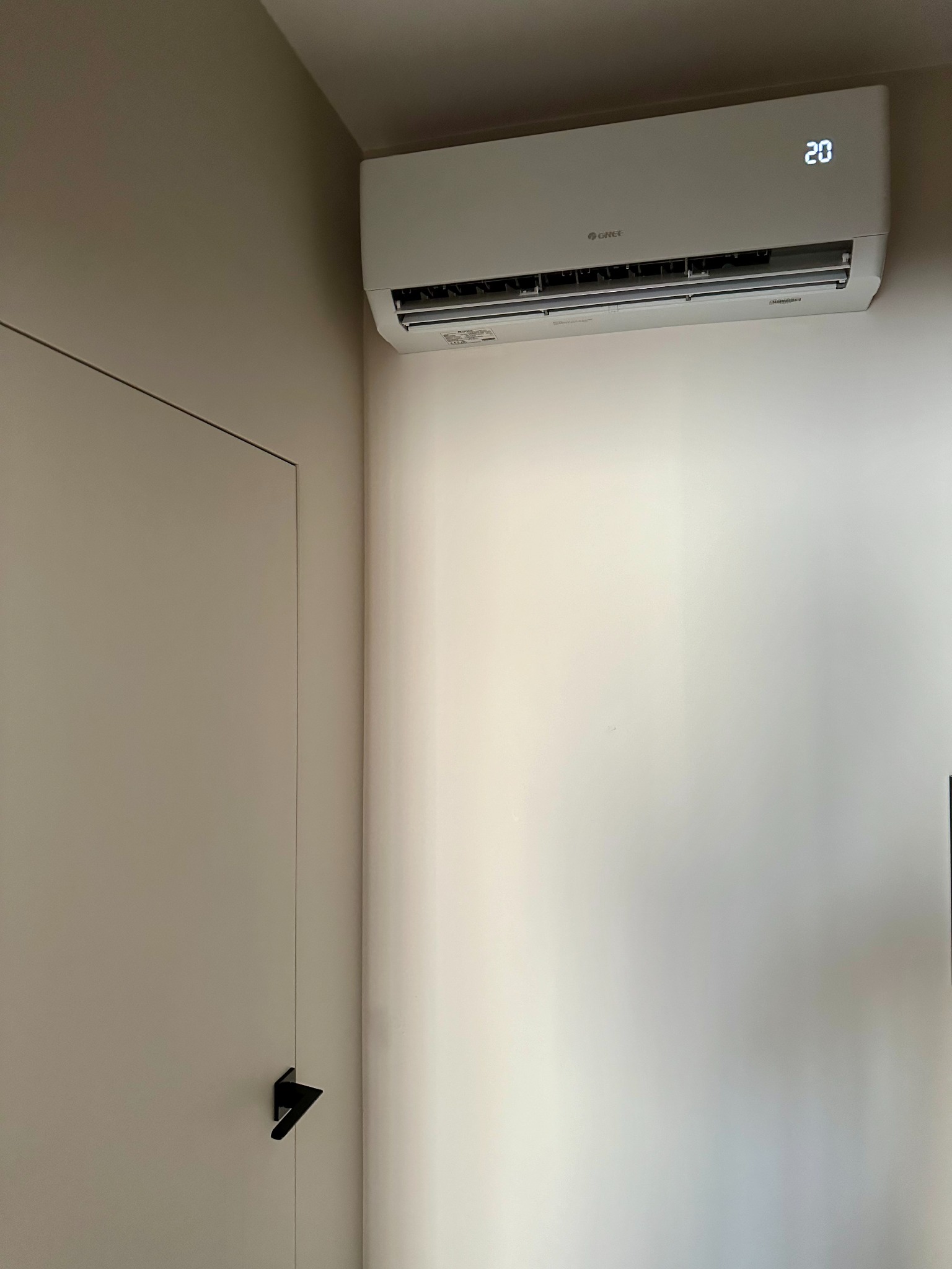 Biały klimatyzator Gree z wyświetlaczem temperatury 20 stopni Celsjusza, zamontowany na ścianie w minimalistycznym wnętrzu obok ukrytych drzwi z czarną klamką.