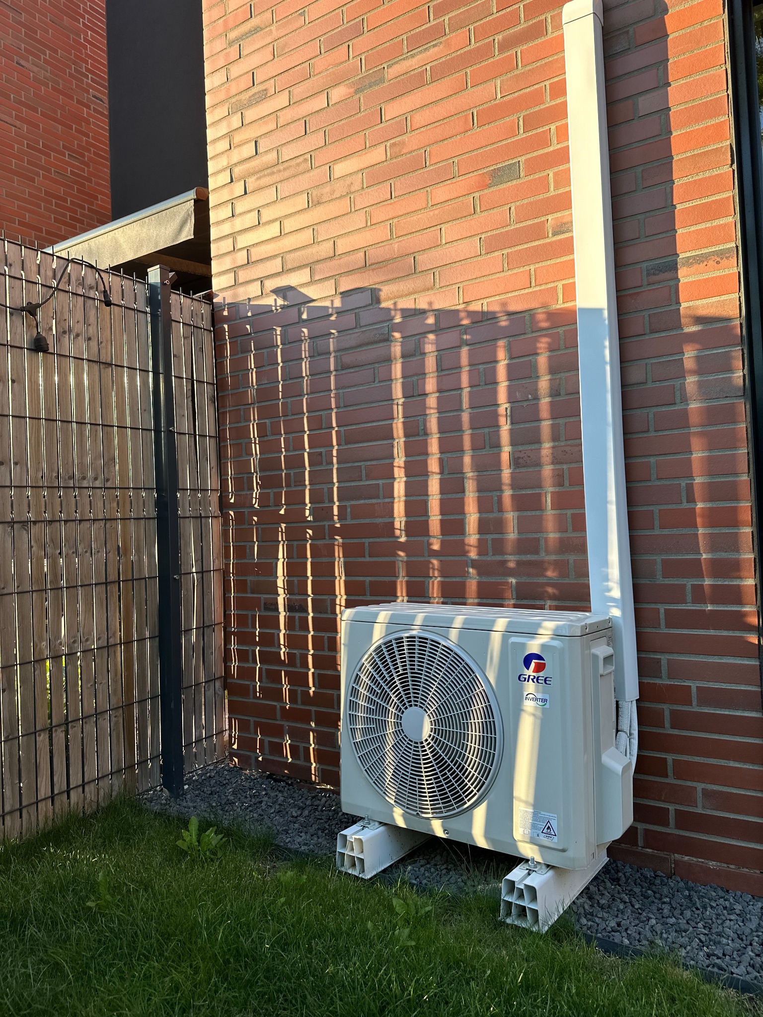 Biała jednostka zewnętrzna klimatyzatora marki Gree, zamontowana na betonowych bloczkach przy ceglanej ścianie domu, z widoczną instalacją odprowadzającą skropliny.