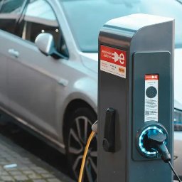 ElektrykWAW.pl Profesjonalny elektryk - Srebrny samochód elektryczny podłączony do stacji ładowania z logo Amsterdam Electric, widoczny kabel zasilający i podświetlony port ładowania.