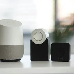 Elektryk.biz Profesjonalny elektryk - Google Home, Nuki Smart Lock i Nuki Bridge na białym blacie, z rozmytym tłem okna i zieleni, prezentacja systemu inteligentnego domu.