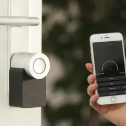 Elektryk.biz Profesjonalny elektryk - Dłoń trzymająca smartfon z aplikacją do sterowania inteligentnym zamkiem na białych drzwiach, widoczny srebrny uchwyt i czarno-srebrny moduł zamka.