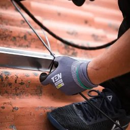 Elektryk.biz Profesjonalny elektryk - Instalator w szarych rękawicach roboczych TEN Protection, w czarnych butach Reebok, mocuje czarny kabel do metalowej szyny na pomarańczowym dachu z dachówki ceramicznej.