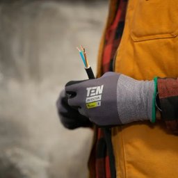 Elektryk.biz Profesjonalny elektryk - Elektryk w rękawicach ochronnych TEN Protection trzyma kabel z trzema żyłami w izolacji niebieskiej, brązowej i żółto-zielonej na tle surowej ściany.