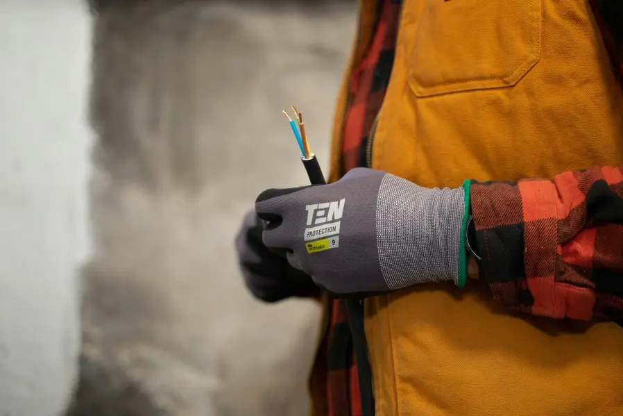Elektryk w rękawicach ochronnych TEN Protection trzyma kabel z trzema żyłami w izolacji niebieskiej, brązowej i żółto-zielonej na tle surowej ściany.