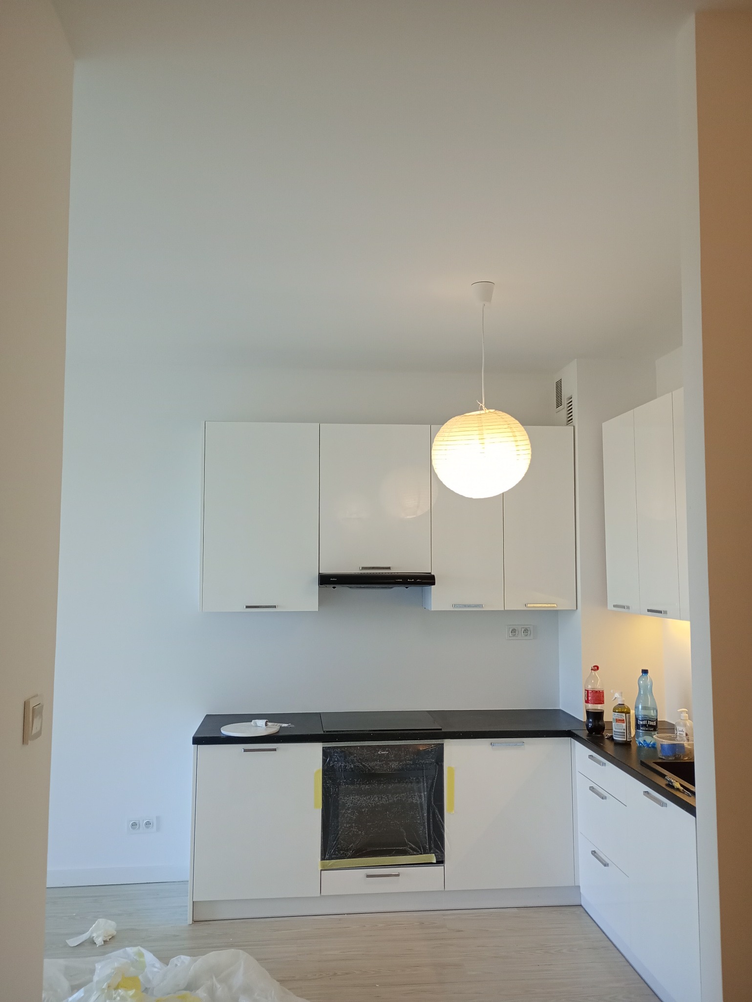 Nowoczesna, jasna kuchnia z białymi szafkami i czarnym blatem, oświetlona papierową lampą. Piekarnik zabezpieczony folią, ściany świeżo pomalowane na biało.