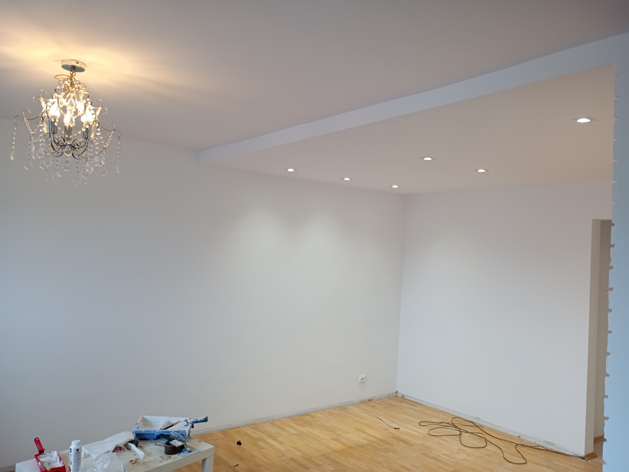 Pomalowane na biało wnętrze z drewnianą podłogą, widoczny kryształowy żyrandol i narzędzia malarskie na stole, w tym kuweta z farbą i wałek.