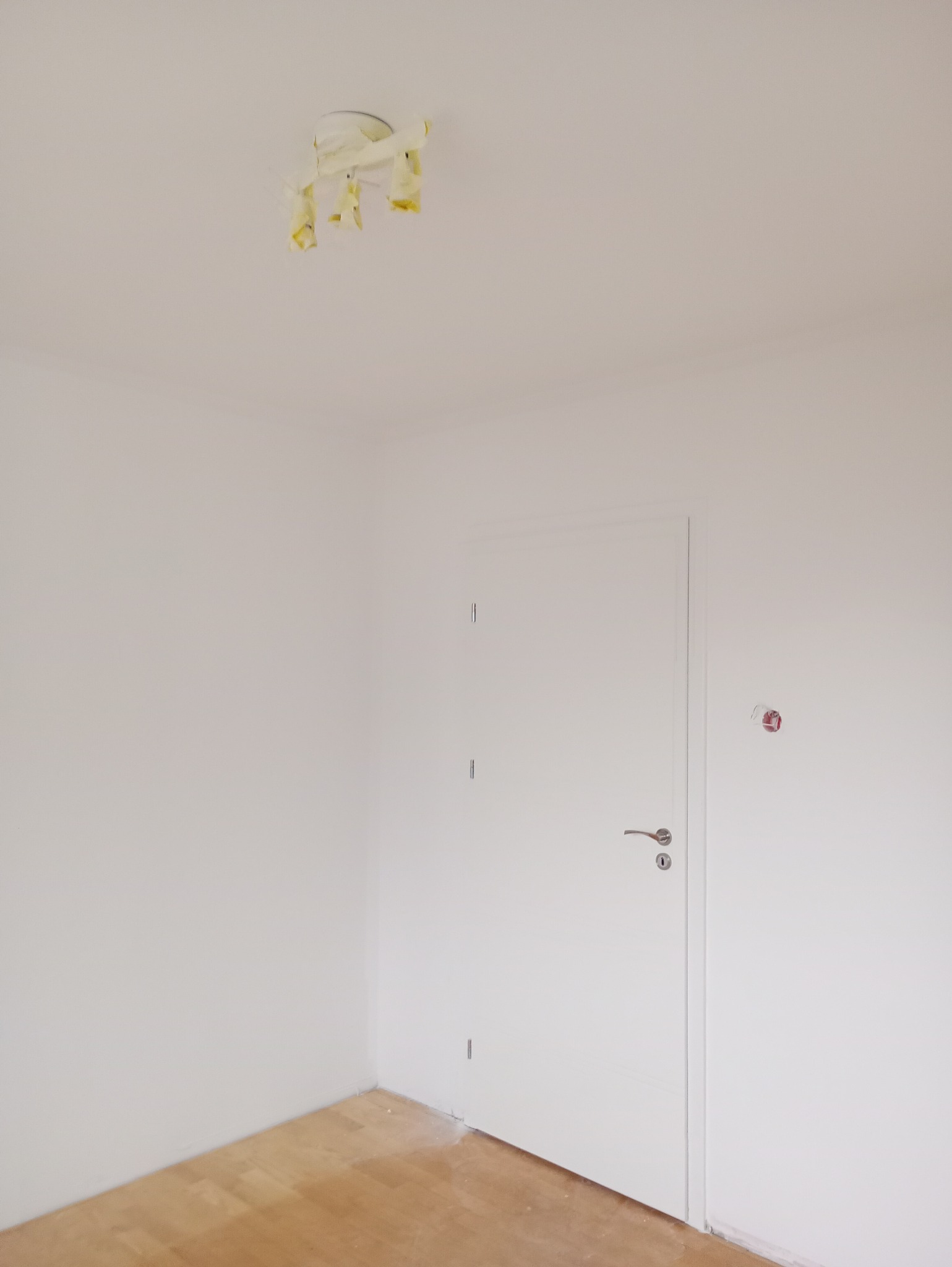 Pomalowane na biało wnętrze pokoju z drewnianą podłogą, białe ściany, białe drzwi. Lampa sufitowa zabezpieczona taśmą malarską.