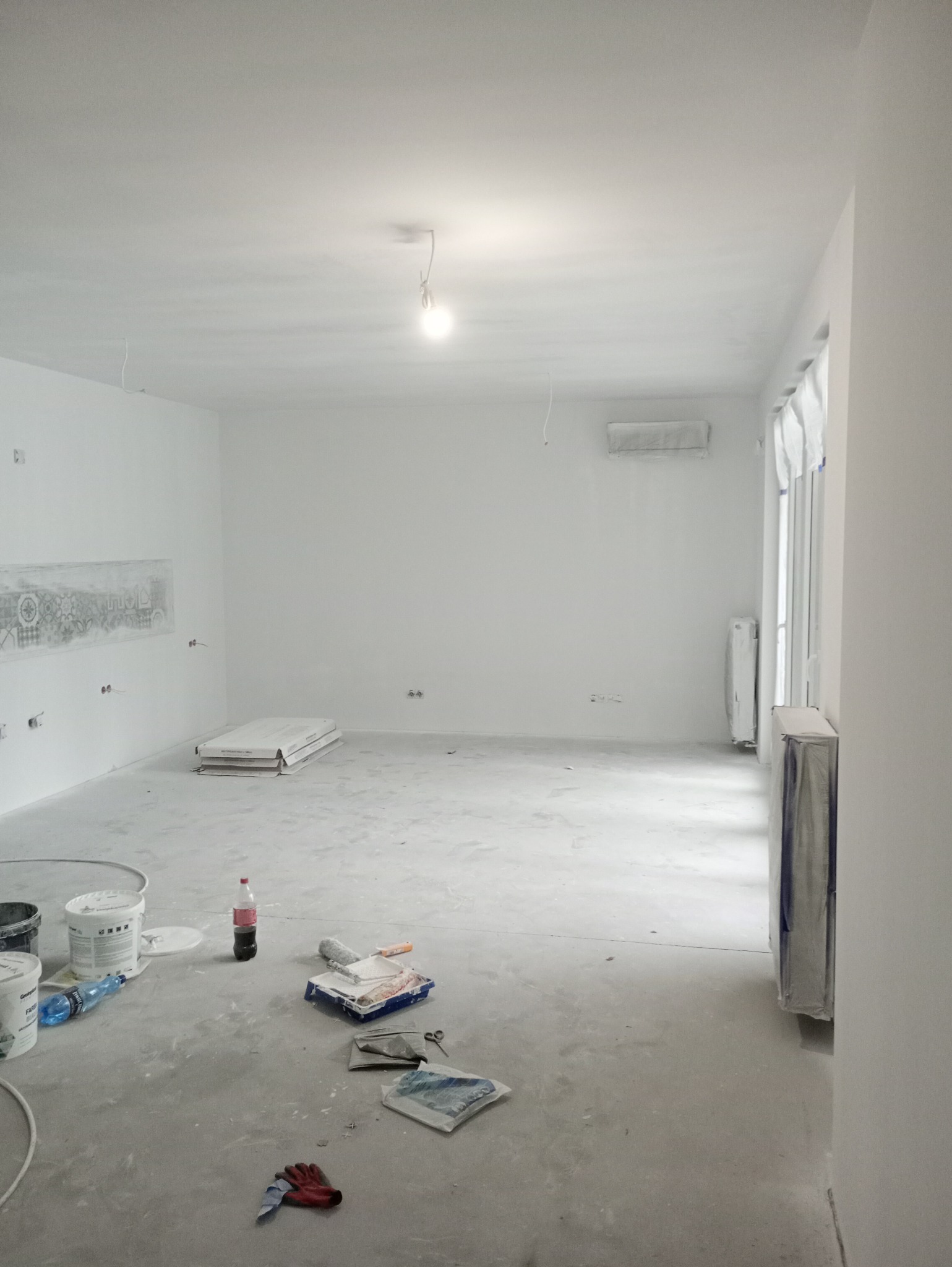 Wnętrze pokoju po malowaniu na biało, widoczne narzędzia malarskie, puszki z farbą, zabezpieczone okno i grzejnik, oraz niedomalowany wzór na ścianie.