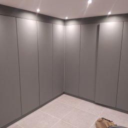 Budowar - Szare, minimalistyczne szafy wnękowe od podłogi do sufitu w rogu pomieszczenia, widoczna torba i odkurzacz na kafelkach podłogowych.