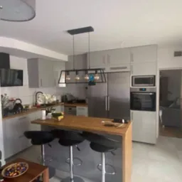 Nowoczesna kuchnia w odcieniach szarości z drewnianym blatem wyspy, czarnymi hokerami i wiszącą lampą z żarówkami Edisona, widok na salon w tle.