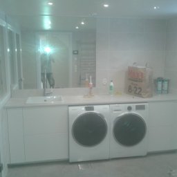 Budowar - Jasne pomieszczenie gospodarcze z dwiema pralkami Samsung pod blatem z wbudowanym zlewem, widoczne płytki na ścianach i lustro z odbiciem osoby robiącej zdjęcie.