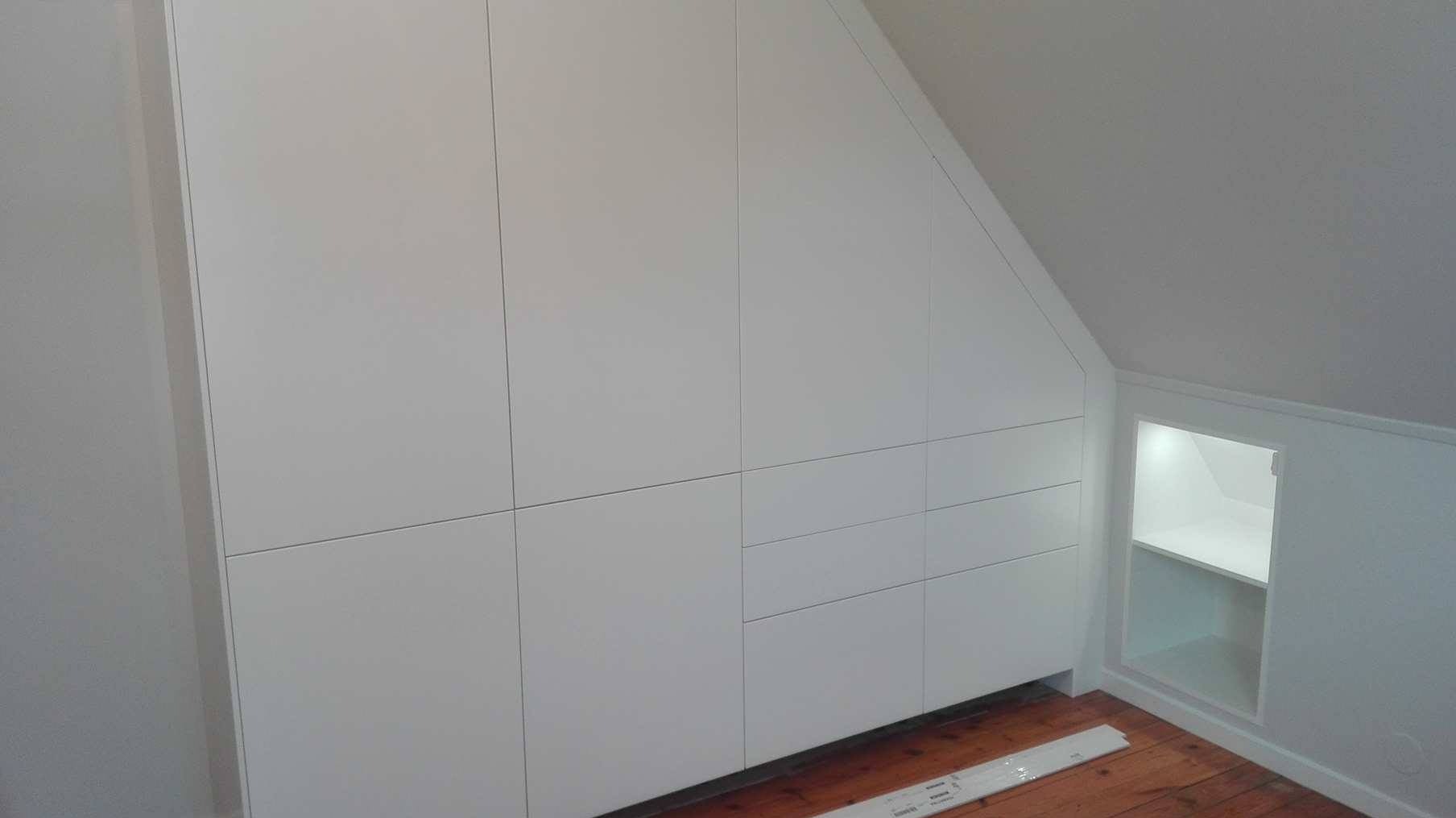 Biała, zabudowana szafa na wymiar, wkomponowana w skos poddasza z dodatkową, oświetloną wnęką z półkami, na podłodze leżą listwy przypodłogowe.