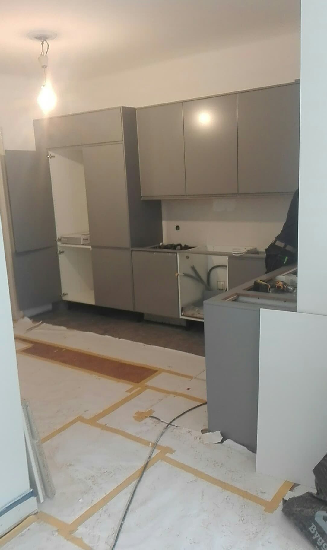 Montaż szarych szafek kuchennych w trakcie prac, podłoga zabezpieczona folią, widoczne narzędzia i kable.