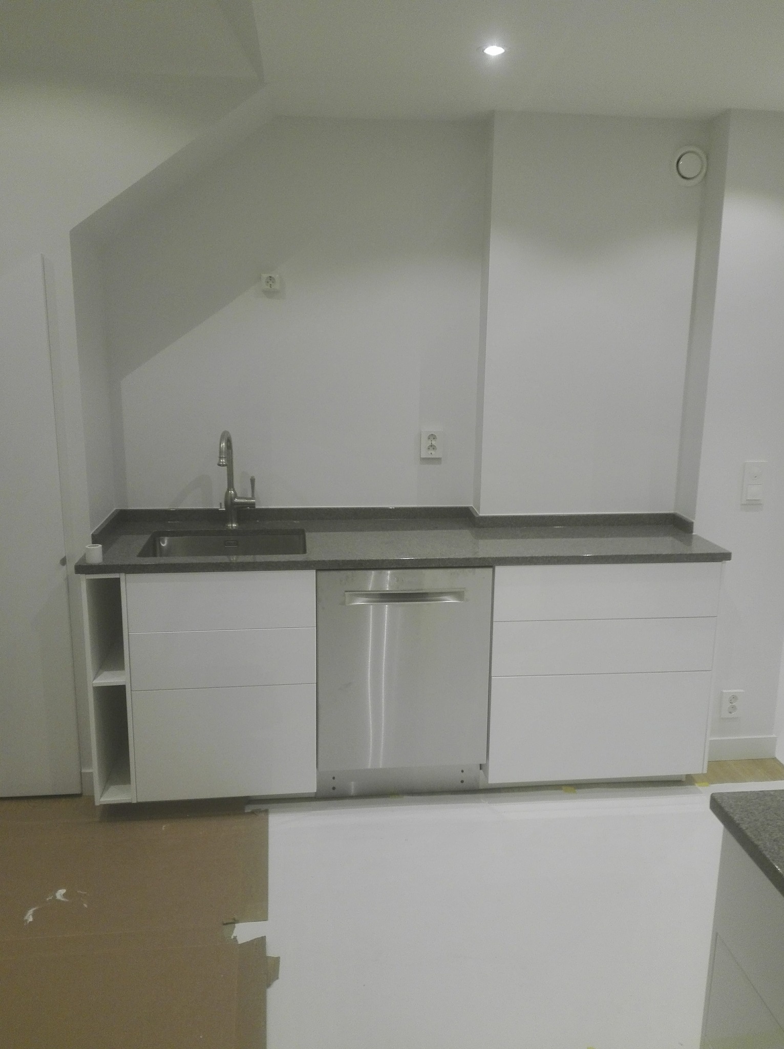 Nowoczesna zabudowa kuchenna z szarym blatem, widoczny zlew, bateria i zmywarka, podłoga zabezpieczona kartonem podczas prac.