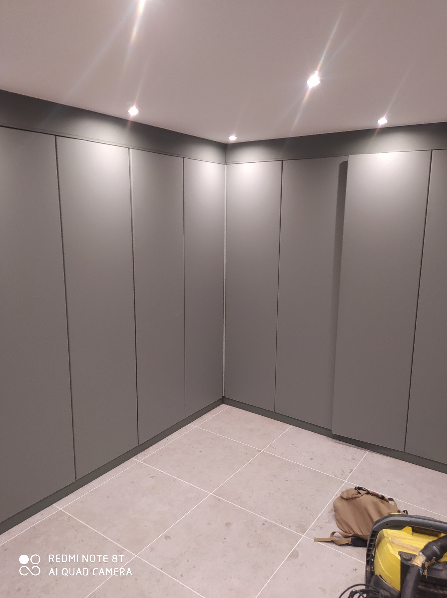 Szare, minimalistyczne szafy wnękowe od podłogi do sufitu w rogu pomieszczenia, widoczna torba i odkurzacz na kafelkach podłogowych.