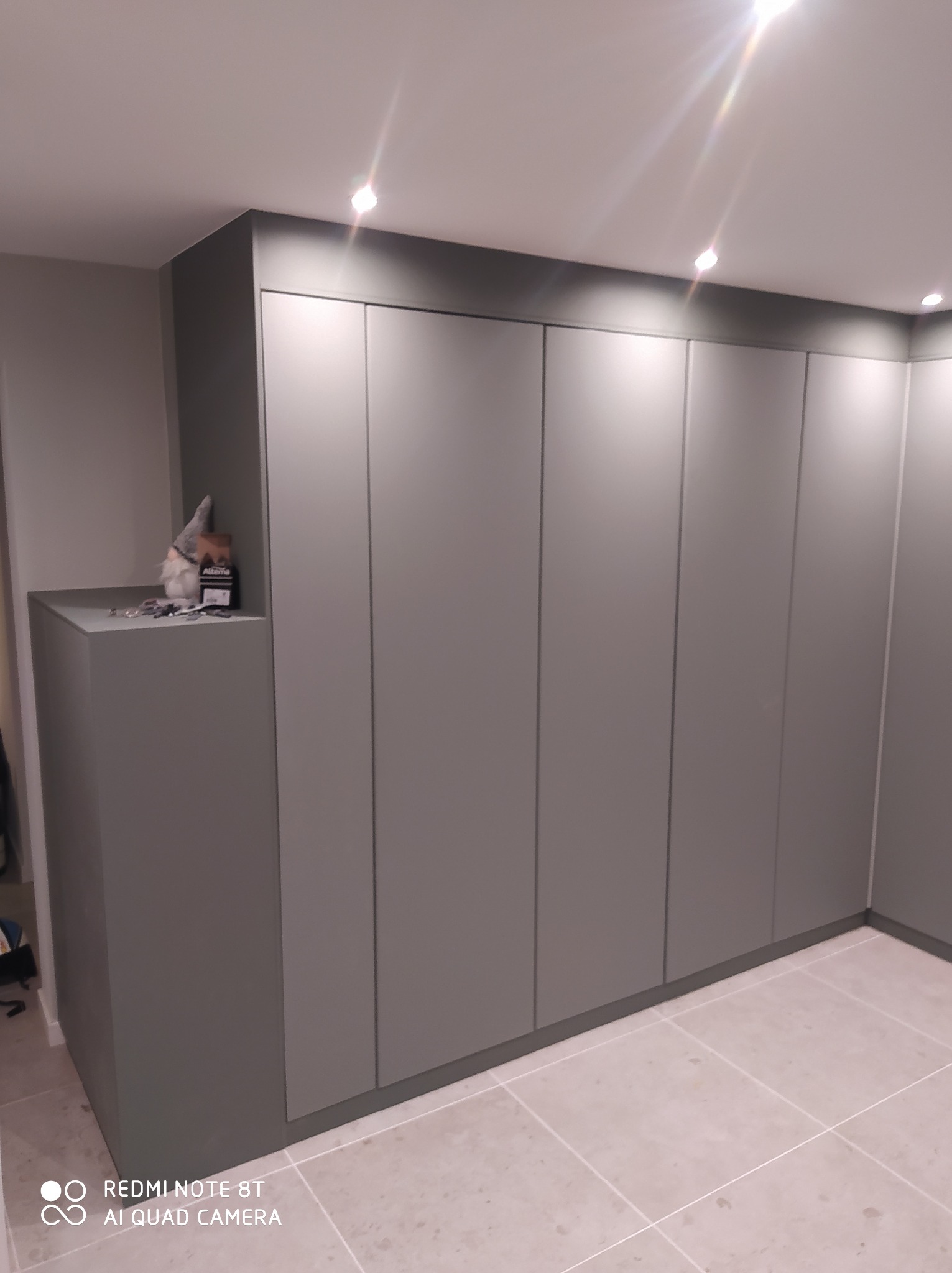 Szara zabudowa szafy wnękowej z oświetleniem punktowym w suficie, na półce dekoracje i akcesoria montażowe.