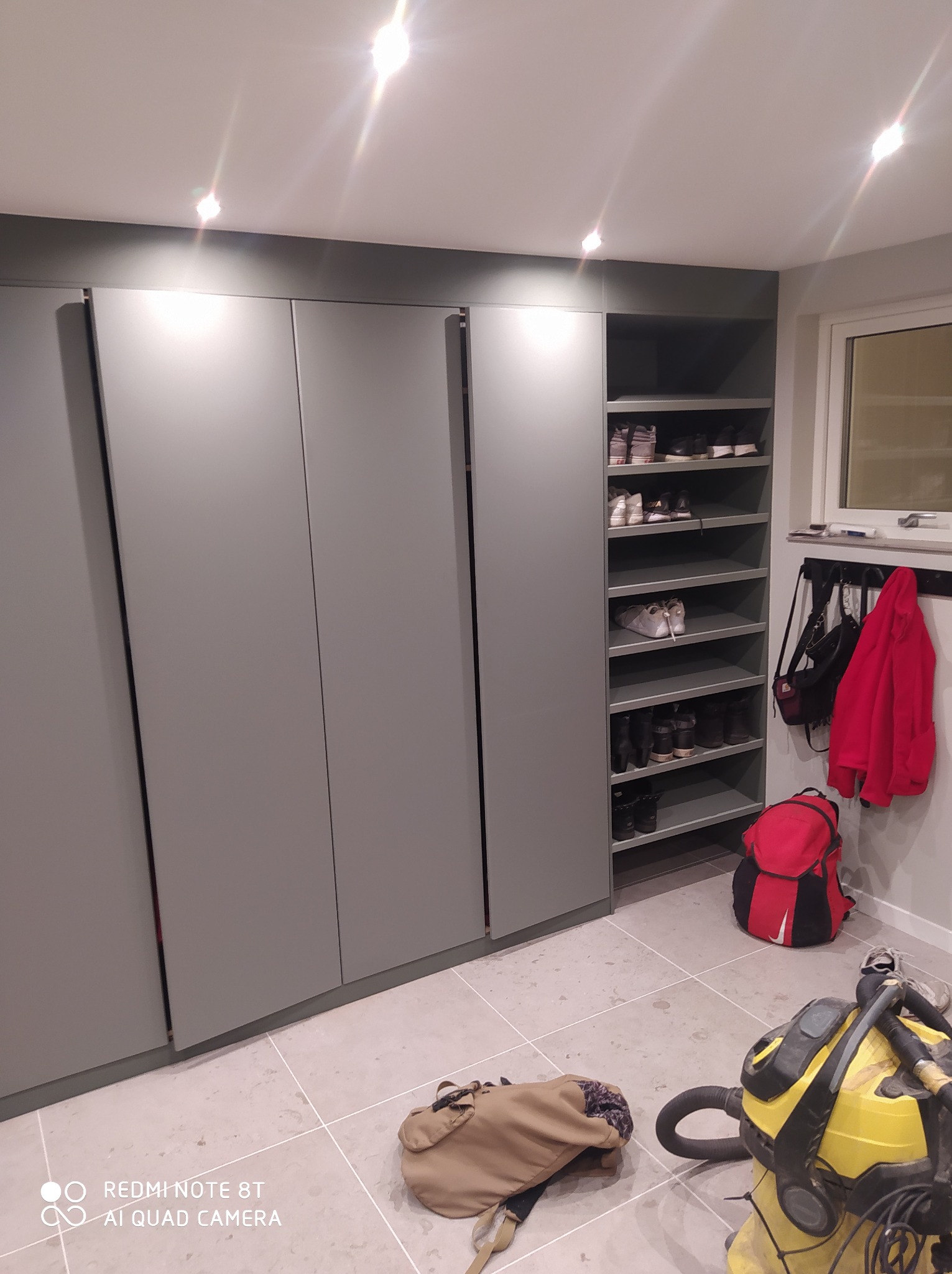 Szara szafa wnękowa z półkami na buty i wieszakiem na ubrania, widoczny odkurzacz i torba na podłodze.