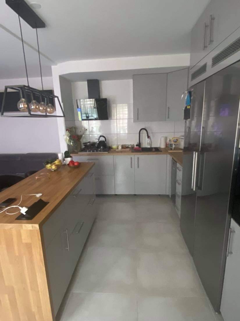 Nowoczesna kuchnia z szarymi szafkami, drewnianym blatem wyspowym z wbudowanymi gniazdkami, czarną armaturą i ozdobną lampą wiszącą z żarówkami Edisona.