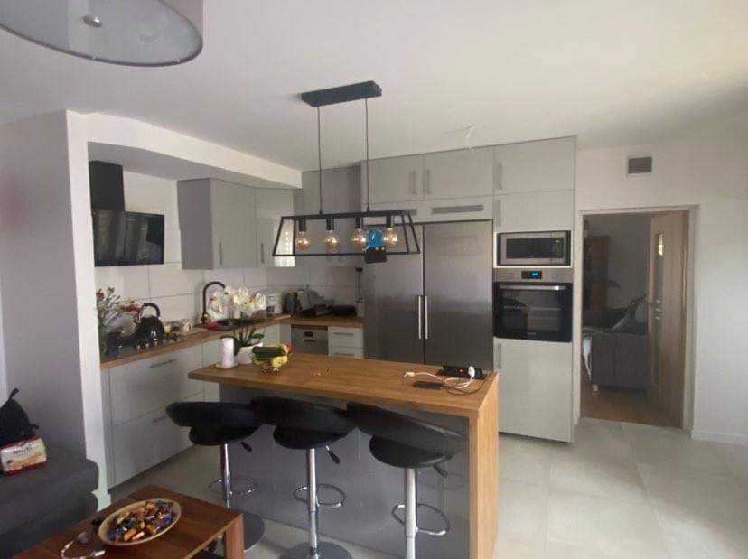 Nowoczesna kuchnia w odcieniach szarości z drewnianym blatem wyspy, czarnymi hokerami i wiszącą lampą z żarówkami Edisona, widok na salon w tle.