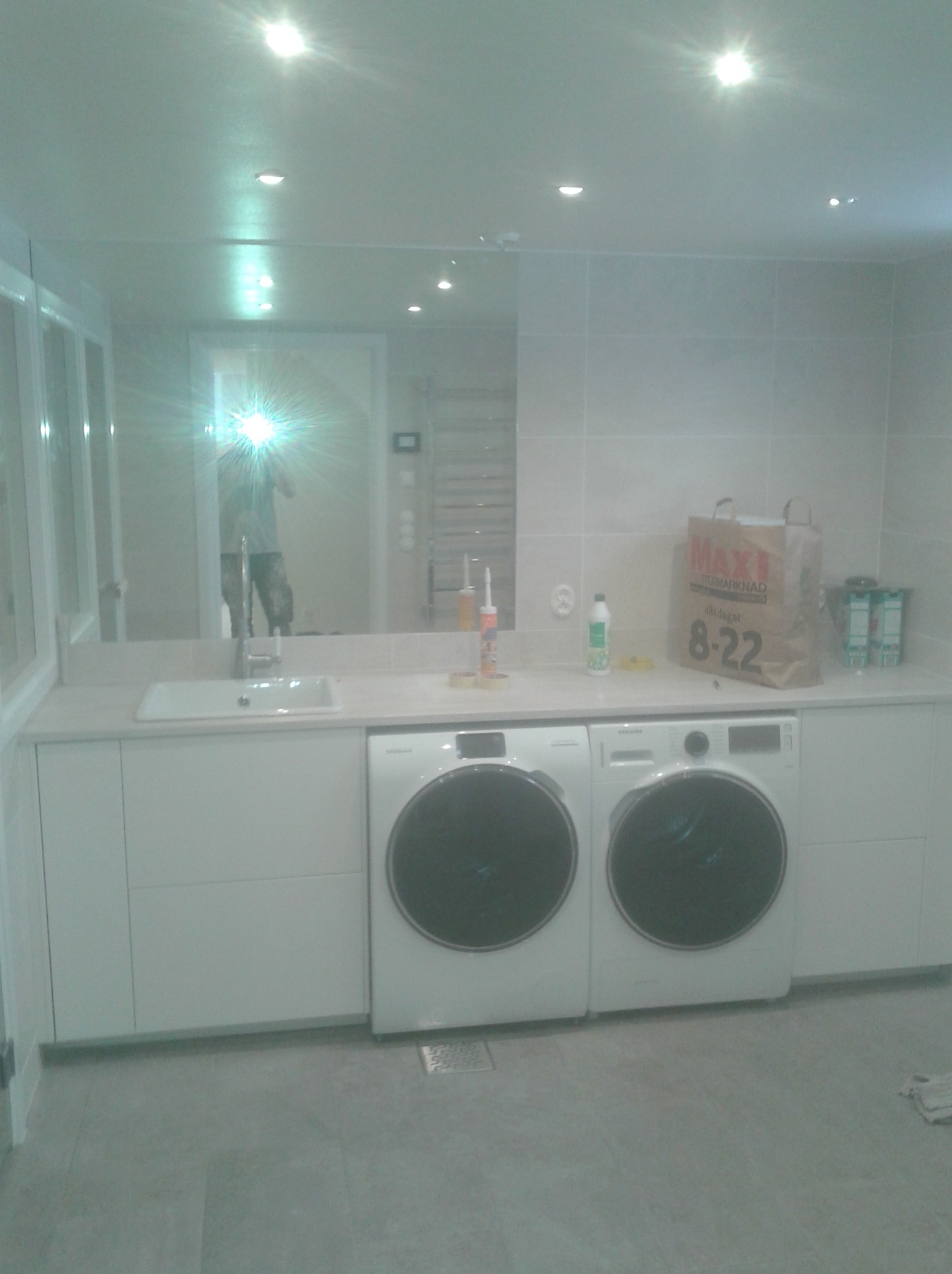 Jasne pomieszczenie gospodarcze z dwiema pralkami Samsung pod blatem z wbudowanym zlewem, widoczne płytki na ścianach i lustro z odbiciem osoby robiącej zdjęcie.