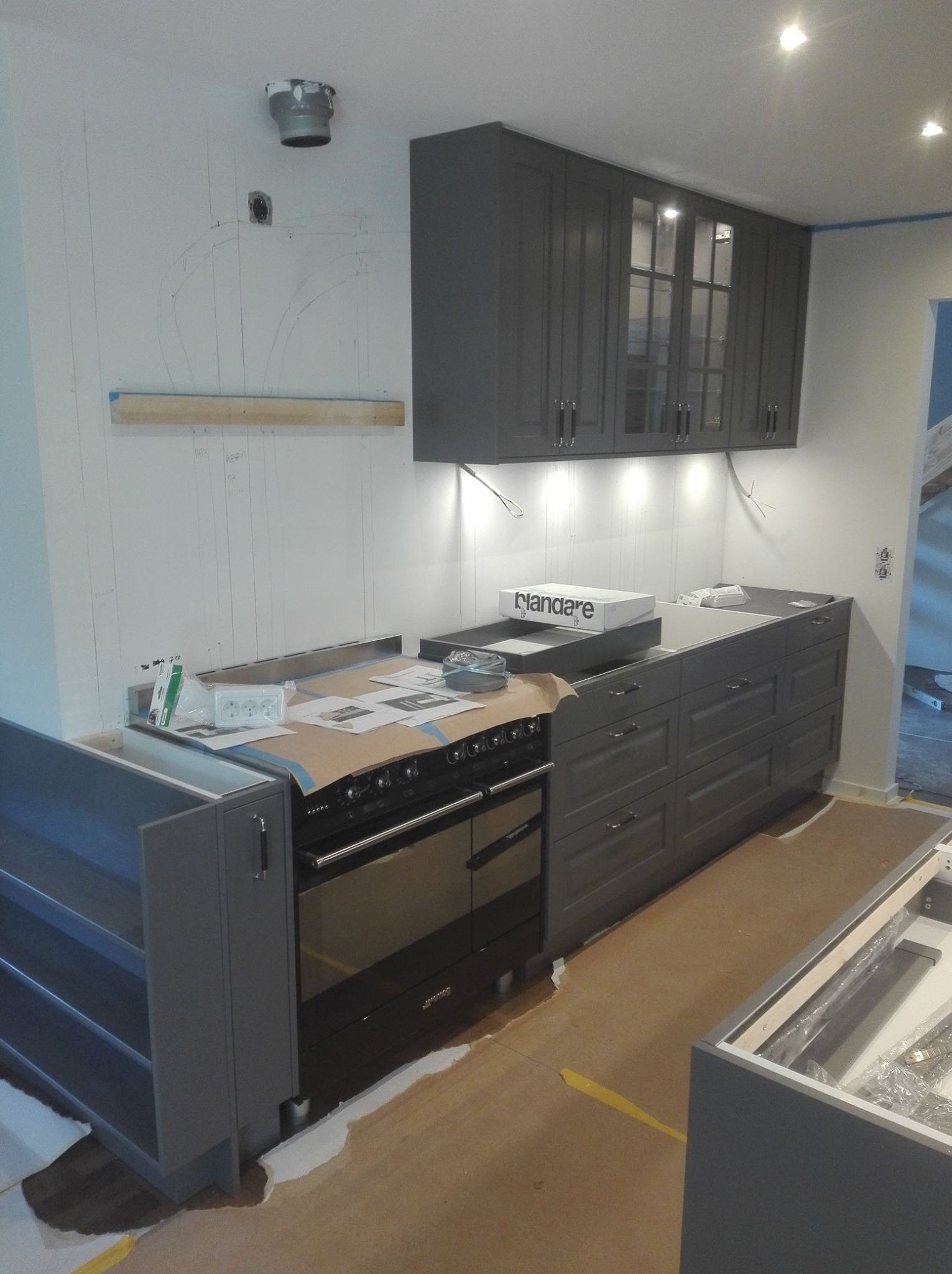 Montaż szarych szafek kuchennych z przeszklonymi frontami, widoczne przygotowania do instalacji oświetlenia podszafkowego, ściana z zaznaczonymi liniami instalacji, zabezpieczona płyta kuchenna.