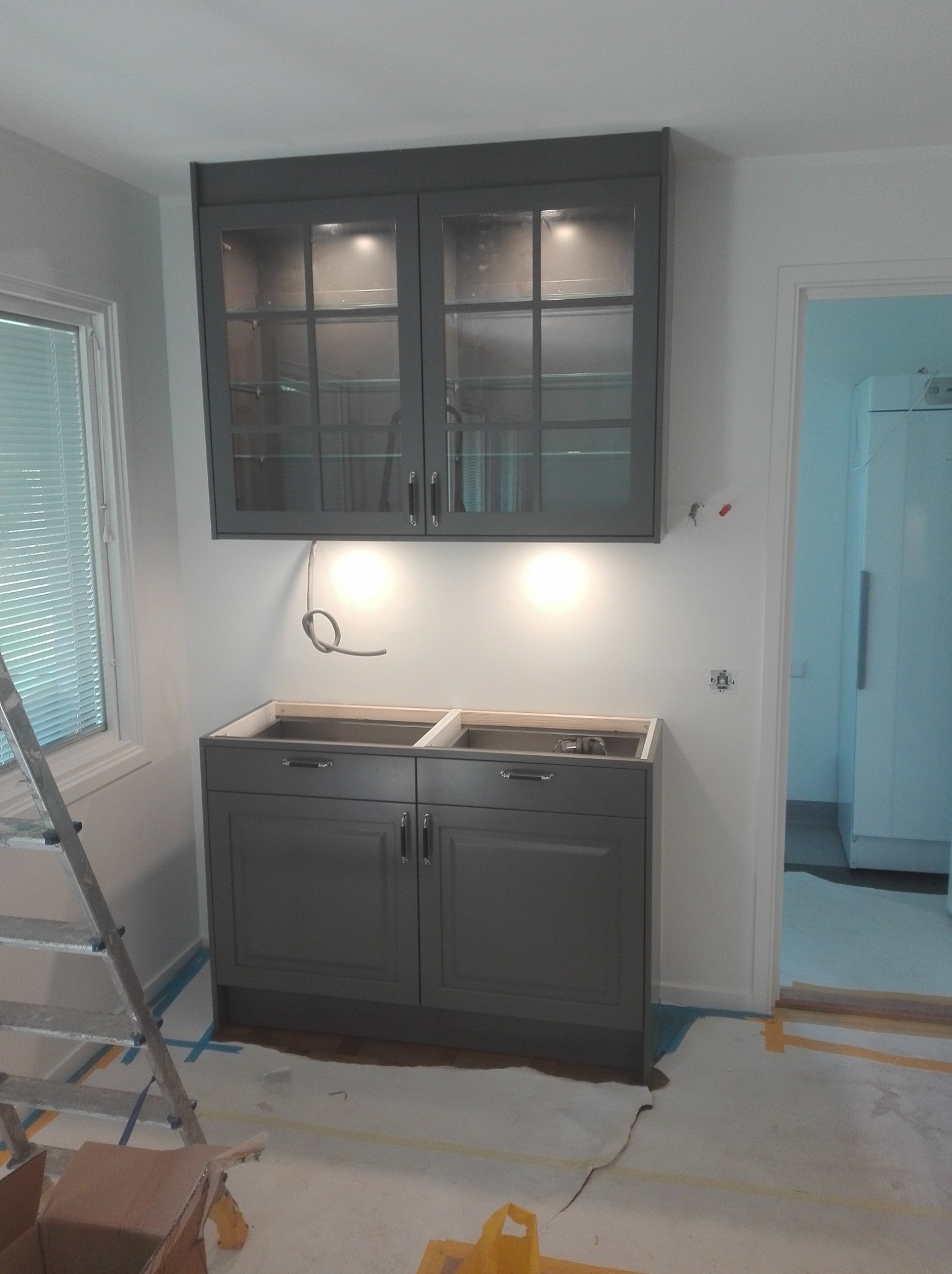Szare szafki kuchenne z przeszklonymi drzwiczkami górnymi, zamontowane bez blatu, z widocznymi przewodami oświetleniowymi i drabina malarska w tle, zabezpieczona podłoga folią malarską.