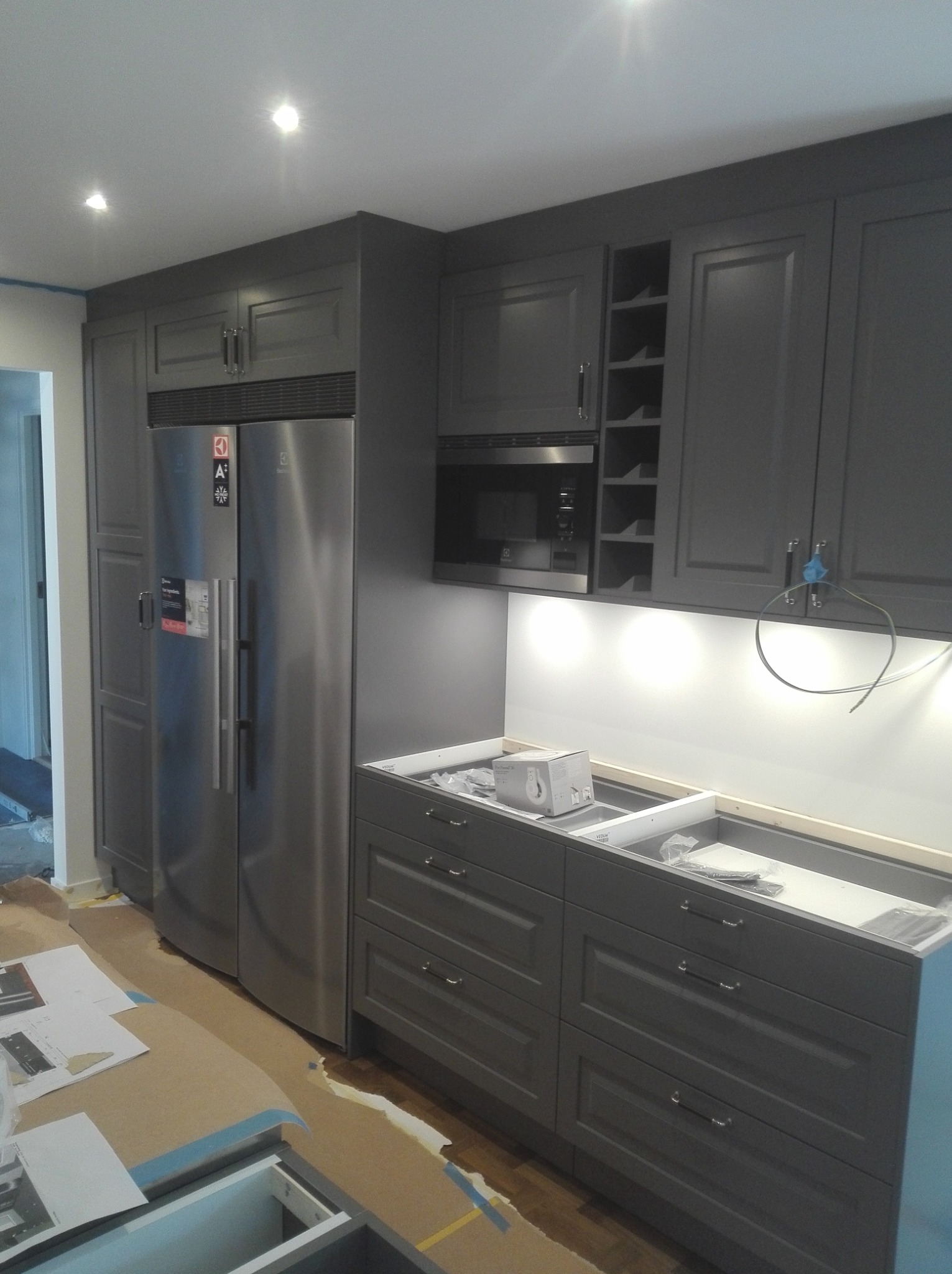 Kuchnia w trakcie montażu: szare szafki kuchenne z srebrnym sprzętem AGD, widoczne elementy instalacji oświetleniowej pod szafkami, zabezpieczona podłoga kartonem.