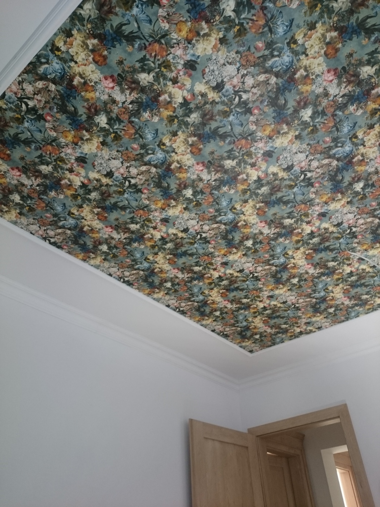 Sufit wykończony wzorzystą tapetą w motywy kwiatowe, widoczne białe listwy sufitowe i fragment ściany z otwartymi drzwiami.